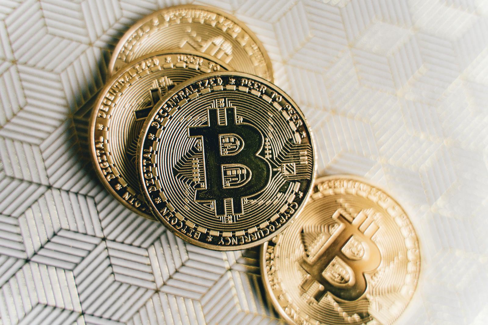 A close-up image of gold Bitcoin cryptocurrency coins on a geometric patterned surface.